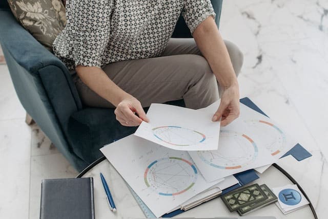Foto de uma mulher sentada com um mapa astral na mão e outros mapas astrais em uma mesinha de centro. Essa foto serve para ilustrar o artigo Astrologia: Entenda o Papel de um Astrólogo do site Ori Mystyco.
